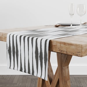 Puede incluir: Un camino de mesa a rayas blanco y gris con un patrón geométrico. El camino de mesa está sobre una mesa de madera con dos copas de vino y un plato.