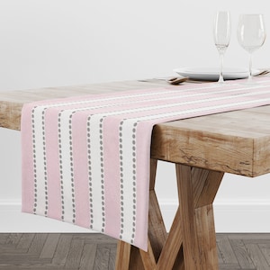 May include: A pink and gray striped table runner with a dotted pattern. The runner is on a wooden table with two wine glasses and a plate.