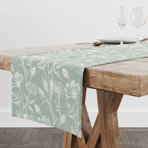 Puede incluir: Un camino de mesa verde salvia con un patrón floral blanco. El camino de mesa está sobre una mesa de madera con dos copas de vino y un plato.
