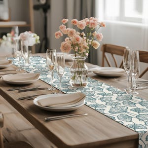 Puede incluir: Un camino de mesa blanco con un patrón de hojas de ginkgo azul. El camino de mesa está colocado sobre una mesa de madera con un jarrón de flores rosas en el centro. Hay dos cubiertos a cada lado del jarrón, cada uno con un plato blanco, cubiertos y una copa de vino.