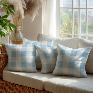 May include: Four square throw pillows with a light blue and white checkered pattern are arranged on a beige sofa. The pillows are in front of a window with natural light. Decorative elements include a vase with dried plants.