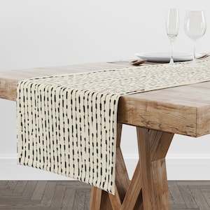 May include: A beige table runner with a black and white geometric pattern. The runner is made of a linen-like fabric and has a subtle sheen.