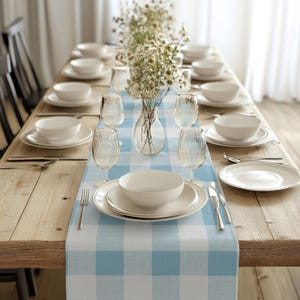 Könnte beinhalten: Ein langer Holztisch, der für ein Essen gedeckt ist. Weiße Teller, Schalen und Gläser sind auf einem blau-weiß karierten Tischläufer angeordnet. Eine klare Glasvase mit Blumen steht in der Mitte. Besteck ist an jedem Platz platziert.