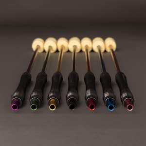 May include: Seven fire poi, each with a different coloured tip: purple, green, gold, black, red, blue, and pink. The handles are black with a textured grip. The wicks are a light beige colour.