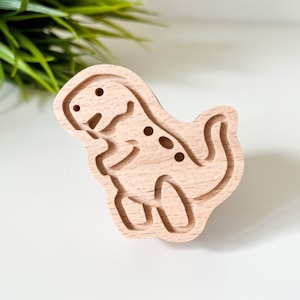 May include: A wooden dinosaur-shaped stamp with a detailed outline of the dinosaur. The stamp is light brown and has a smooth surface.