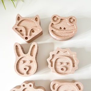 Könnte beinhalten: Sechs Holztiere mit verschiedenen Tiergesichtern, die in sie geschnitzt sind. Die Tiere sind ein Fuchs, ein Frosch, ein Kaninchen, ein Löwe, eine Biene und ein Bär.