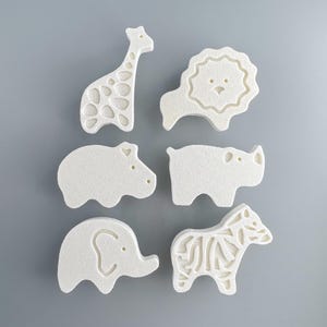 Peut inclure: Ensemble de six jouets de bain blancs en forme d'animaux différents : une girafe, un lion, un hippopotame, un rhinocéros, un éléphant et un zèbre. Les jouets sont faits d'un matériau doux et texturé.