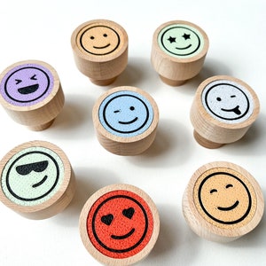 Könnte beinhalten: Eine Sammlung von Holzschrankknöpfen mit verschiedenen Emoji-Gesichtern. Jeder Knopf zeigt einen anderen Ausdruck, darunter lächeln, zwinkern und Sonnenbrillen tragen. Die Emoji-Gesichter sind in verschiedenen Farben wie Gelb, Blau und Rot gehalten.