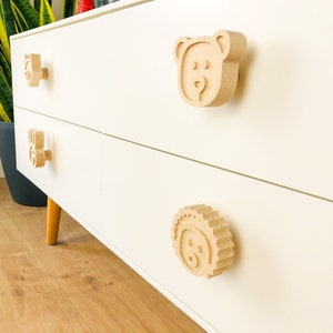 Puede incluir: Cómoda blanca con tres cajones con pomos de madera con forma de oso, conejo y erizo. Los pomos son de color marrón claro.
