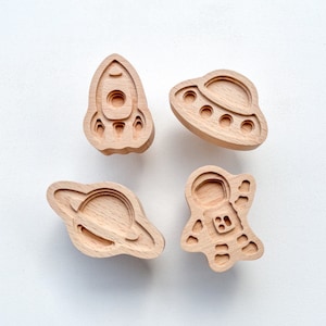 May include: Four wooden space-themed cutouts: a rocket, a UFO, a planet with rings, and an astronaut. The cutouts are light brown and have a layered design, creating a 3D effect. The objects are arranged on a white background.