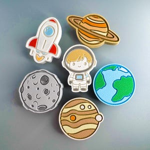 May include: A set of six space-themed 3D printed ornaments. The ornaments include a rocket, an astronaut, a moon, a planet with rings, a planet with stripes, and a planet with a green and blue surface.