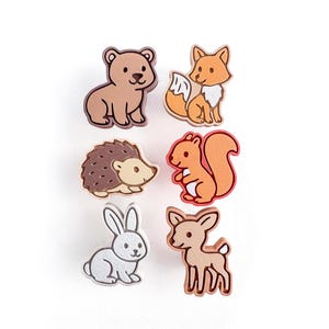 Könnte beinhalten: Set aus sechs Holz-Tierausschnitten: ein Bär, ein Fuchs, ein Igel, ein Eichhörnchen, ein Kaninchen und ein Reh. Jeder Ausschnitt ist mit detaillierten Merkmalen bemalt und in einer dunkleren Farbe umrandet. Der Bär ist braun, der Fuchs ist orange und das Eichhörnchen ist ebenfalls orange.