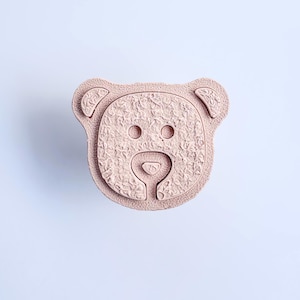 Könnte beinhalten: Ein hellrosa Knopf in Bärenform mit einer strukturierten, fellartigen Oberfläche. Das Design zeigt zwei kreisförmige Augen, eine kleine dreieckige Nase und einen abgerundeten Schnauzenbereich, die alle in die Bärenkopf-Silhouette mit zwei kleinen Ohren integriert sind.