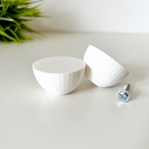 Op de afbeelding: Twee witte, halfronde houten kastknoppen met verticale groeven. Een zilveren schroef is in de buurt geplaatst. De knoppen hebben een platte bovenkant en een afgeronde, komvormige vorm. De knoppen zijn waarschijnlijk voor meubels.