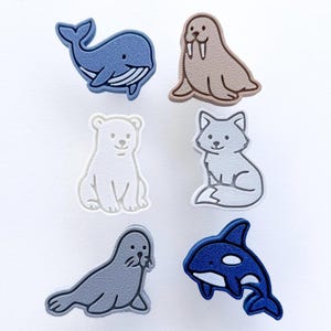 Peut inclure: Un ensemble de breloques d'animaux de dessin animé colorés. L'ensemble comprend une baleine bleue, un morse beige, un ours polaire blanc, un renard gris, un phoque gris et un orque bleu et blanc. Ces breloques sont probablement décoratives.