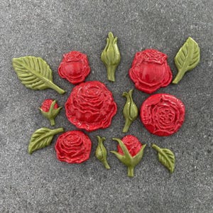 May include: A selection of red rose blossoms, buds, and green leaves arranged on a grey surface. The roses display detailed petal textures. The leaves and buds are a contrasting green.