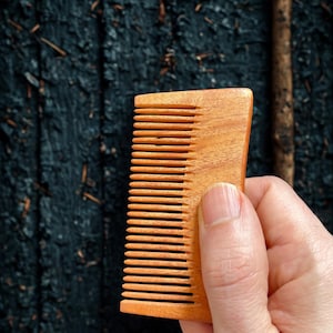 Könnte beinhalten: Nahaufnahme eines Holzkamms mit feinen Zähnen. Der Kamm wird in einer Hand vor einem dunklen, strukturierten Hintergrund gehalten.