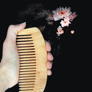 May include: A wooden comb with a rounded edge and fine teeth, held in a hand against a black background with pink flowers.