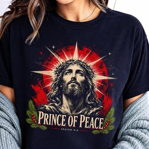May include: A black t-shirt featuring a graphic of a bearded figure wearing a crown of thorns, set against a red sunburst background. Below the figure, the text reads "PRINCE OF PEACE" and "ISAIAH 9:6", framed by green pine branches and red berries.