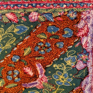 May include: A close-up of a batik fabric with a colorful floral pattern. The fabric is green, orange, blue, yellow, and pink. The fabric has a border with a repeating pattern of triangles and circles. The text "Batik Tulis" is visible in the upper right corner of the image.