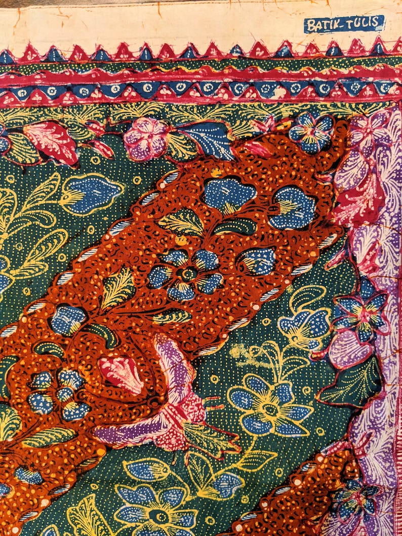 May include: A close-up of a batik fabric with a colorful floral pattern. The fabric is green, orange, blue, yellow, and pink. The fabric has a border with a repeating pattern of triangles and circles. The text "Batik Tulis" is visible in the upper right corner of the image.