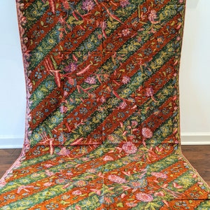 May include: A brown and green batik fabric with a floral pattern. The fabric has a diagonal stripe pattern with a repeating floral motif. The fabric is draped over a white surface.