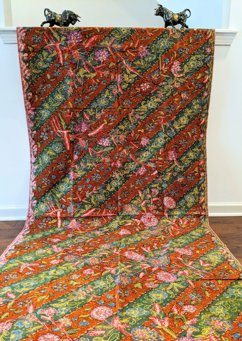 May include: A brown and green batik fabric with a floral pattern. The fabric has a diagonal stripe pattern with a repeating floral motif. The fabric is draped over a white surface.