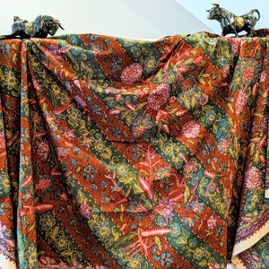 May include: A brown, green, and gold patterned fabric with a diagonal stripe design. The fabric is draped over a surface with two small black bull figurines on top.