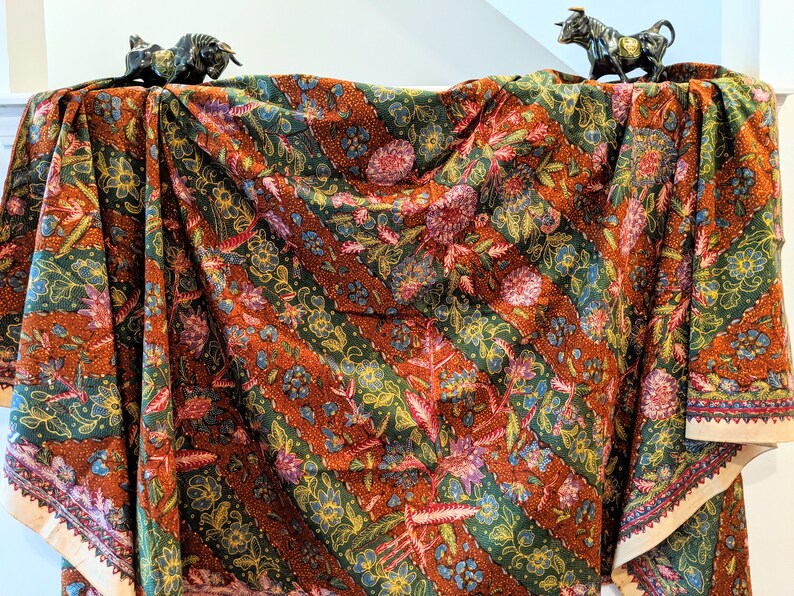 May include: A brown, green, and gold patterned fabric with a diagonal stripe design. The fabric is draped over a surface with two small black bull figurines on top.