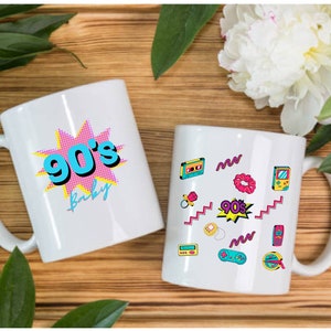 May include: Two white ceramic mugs with 90's themed designs. One mug features a pink and blue graphic with the text "90's Baby". The other mug has a white background with a variety of 90's themed graphics, including a cassette tape, a scrunchie, a game console, and a pacifier.