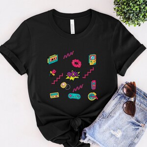 May include: Black t-shirt with a 90s theme featuring a variety of colorful retro icons, including a cassette tape, a scrunchie, a game console, and a cell phone. The text "90's" is printed in a pink and yellow starburst.