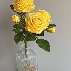 May include: A bouquet of yellow roses in a clear glass vase with a textured surface. The vase is filled with small white stones.
