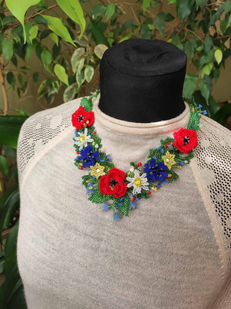 May include: A beaded necklace with a floral design. The necklace features red, blue, white, and yellow flowers with green leaves. The necklace is on a black mannequin.