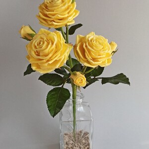 May include: A bouquet of yellow roses in a clear glass vase filled with small stones. The roses have a soft, velvety texture and are arranged in a simple, elegant style.