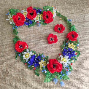 May include: A beaded necklace and earrings set featuring red poppies, white daisies, blue forget-me-nots, and green leaves. The necklace is a floral garland design.