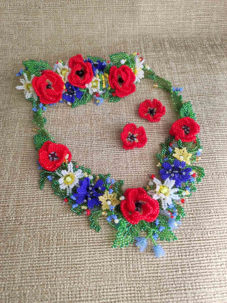 May include: A beaded necklace and earrings set featuring red poppies, white daisies, blue forget-me-nots, and green leaves. The necklace is a floral garland design.