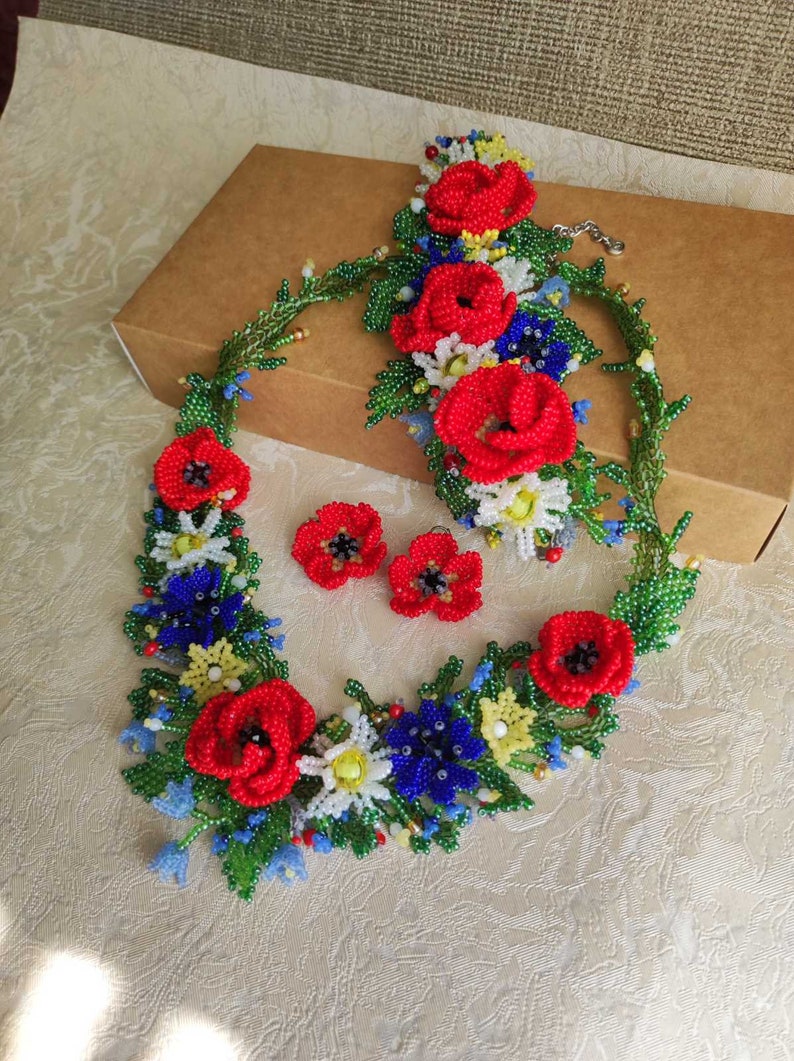 May include: A beaded flower necklace and earrings set with red poppies, white daisies, blue forget-me-nots, and green leaves.