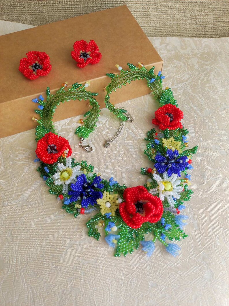 May include: A beaded necklace with a floral design featuring red poppies, blue and white flowers, and green leaves. The necklace is accompanied by a pair of matching earrings.