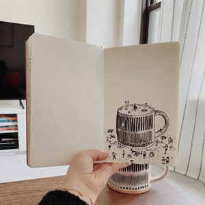 May include: An open notebook with a black and white ink drawing of a large coffee mug with a beach scene inside. The mug is sitting on a beach with people and umbrellas. The notebook is sitting on a wooden table.