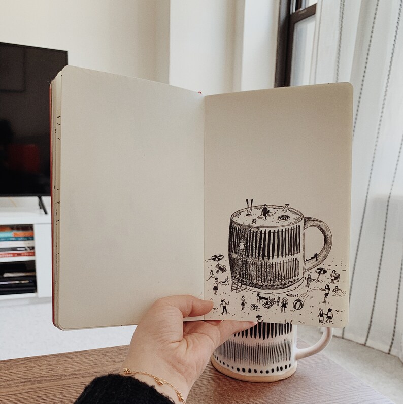May include: An open notebook with a black and white ink drawing of a large coffee mug with a beach scene inside. The mug is sitting on a beach with people and umbrellas. The notebook is sitting on a wooden table.