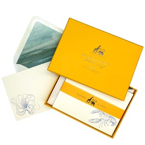 May include: A yellow box with a silver logo that says "M&G Stationery for the discerning" and "Special Quality Stationery London". The box is open and shows a stack of white stationery with a blue octopus illustration and a stack of white stationery with a blue lobster illustration. The stationery is labeled "Mustard & Gray".