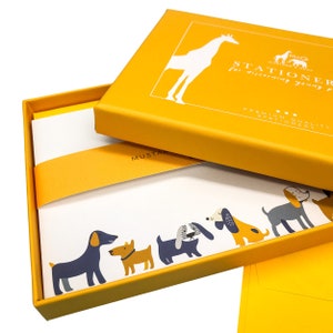 May include: A yellow box with a white giraffe illustration and the text "Stationer for discerning young people" and "Premium Quality Stationery". The box is open and shows a stack of white note cards with a border of colorful dogs.