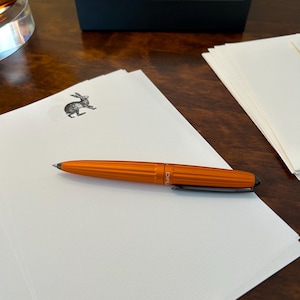 May include: A metallic orange ballpoint pen with a ribbed barrel and a black clip resting on a stack of white stationery. The top sheet features a small black ink illustration of a rabbit. A black Diplomat brand box is positioned in the background.