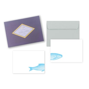 May include: A set of note cards with matching envelopes. The cards are white with a blue fish illustration. The set includes a purple box with gold accents and the words "Note Cards" printed on the front.