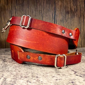 May include: A brown leather camera strap with silver hardware and a metal clip. The strap is folded in half and the ends are overlapping.