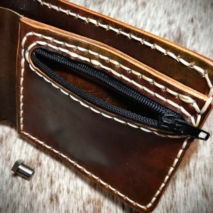 May include: A brown leather wallet with a zippered pocket and white stitching. The wallet is open and the zipper is pulled down to reveal the pocket.