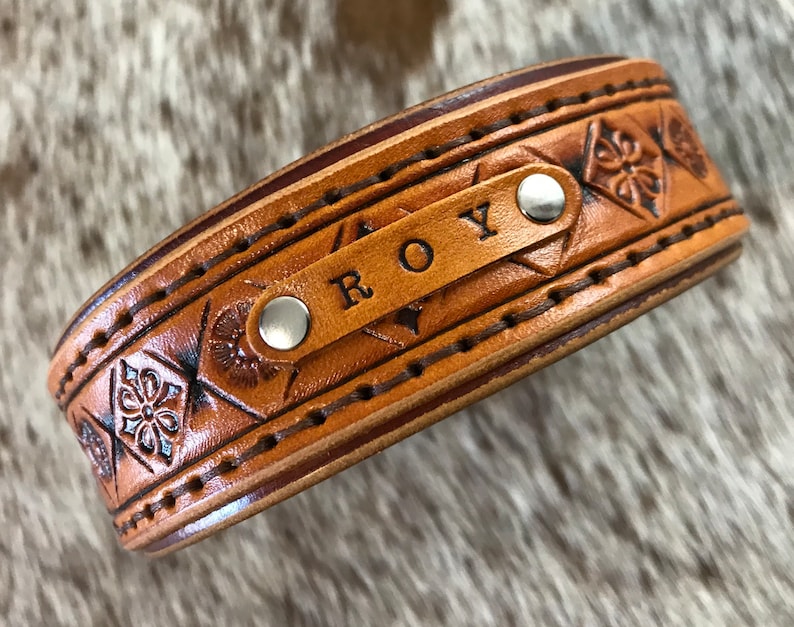 Puede incluir: Un brazalete de cuero marr&oacute;n con un patr&oacute;n geom&eacute;trico y el nombre "ROY" grabado en &eacute;l.