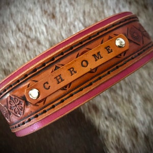 Puede incluir: Un brazalete de cuero marr&oacute;n con un borde rosa y un patr&oacute;n floral decorativo. El brazalete tiene un remache plateado y el nombre "CHROME" est&aacute; grabado en la parte delantera.