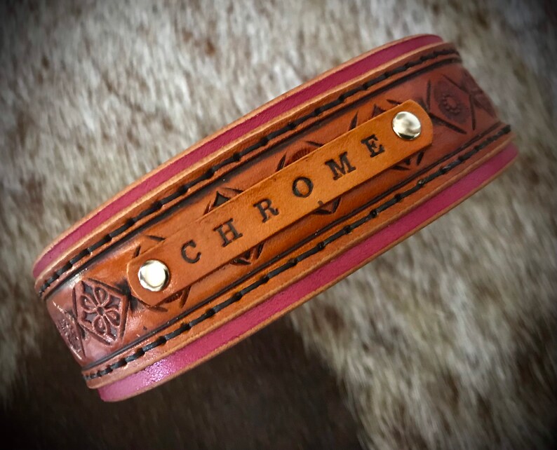 Puede incluir: Un brazalete de cuero marr&oacute;n con un borde rosa y un patr&oacute;n floral decorativo. El brazalete tiene un remache plateado y el nombre "CHROME" est&aacute; grabado en la parte delantera.