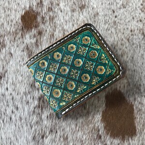 May include: A teal leather wallet with a gold and green geometric pattern. The wallet has a stitched edge and is closed.
